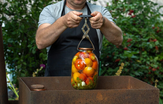 A person is using a tool to secure a jar filled with colorful vegetables. The activity takes place in a garden surrounded by greenery and plants during the warm weather