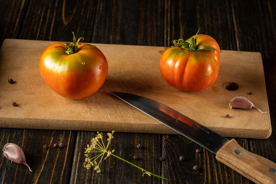 Two ripe tomatoes sit on a wooden cutting board next to garlic cloves. A knife lies beside them as the kitchen scene shows ready ingredients for cooking