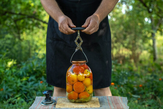A person holds a jar lifter, lifting a jar filled with tomatoes. The scene takes place in a garden with green plants and trees in the background during daylight