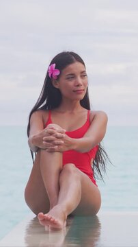 Happy Asian beauty swimwear model wearing red laced crisscross one piece swimsuit sitting by the infinity pool relaxing enjoying summer travel vacations on luxury ocean destination.