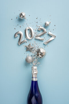 Happy New Year 2027 celebration with champagne bottle, silver confetti and festive ornaments on vertical pastel blue background