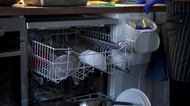 Person wearing gloves loading dishes and glasses into dishwasher machine at home