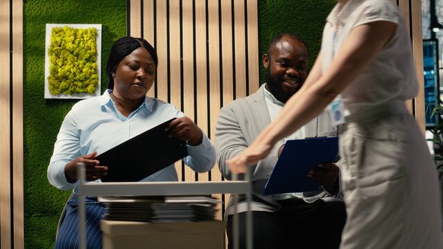 Businesswoman secretary walks with a trolley cart and gives files and corporate registration forms to people sitting in a hallway line while candidates prepare for career opportunities.