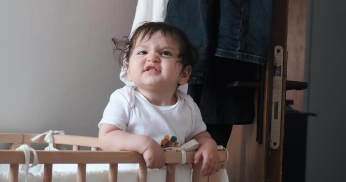 Funny toddler girl standing in wooden crib making grumpy faces then wrinkling nose with a cheeky smile