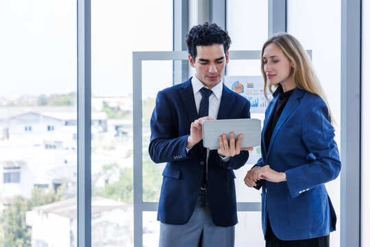 Professional businesswoman and businessman discussing data on a digital tablet near a glass presentation board in a modern office environment.