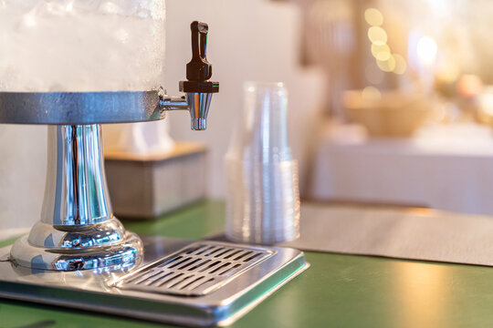 Close-up view of a beverage dispenser filled with cold iced water or juice, featuring a metallic chrome stand and spigot Stacked clear plastic cup are placed in background, event, or cafe setup.