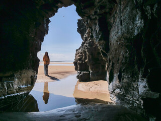 Person stands in cave entrance, gazing out to beach. Warm sunlight highlights wet sand and calm water. Reflection shows peaceful solitude. Perfect for travel, adventure, or nature themes © mark_gusev