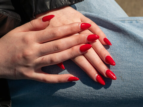 Close up of hands with bright red nails resting on denim. Fingers gently clasped, showing relaxed posture. Sunlight highlights texture of skin and fabric. Image conveys calmness, personal care