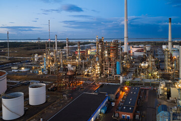 Naklejka premium Large Oil Refinery Facility Illuminated at Twilight Featuring Tall Chimneys and Storage Tanks