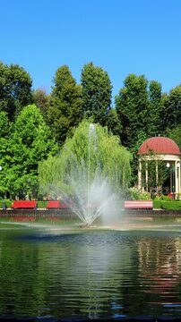 A fountain on the water in a picturesque city garden with a gazebo. A beautiful pond in a city park in Kharkiv on a sunny summer day. Splashes and jets of water from the floating fountain