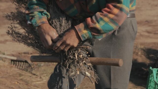 Rural craftsman using a wooden lever tool to tightly compress and wire bind a traditional twig broom outdoors