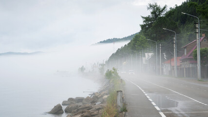 Fototapeta premium Thick fog over a coastal road with driving cars at Lake Baikal, Russia. Low visibility weather conditions on a lakeside highway with houses and streetlights.
