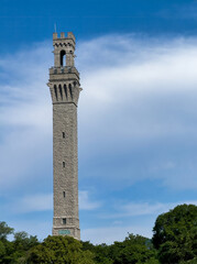 Fototapeta premium Provincetown, Massachusetts, USA - June 24, 2026: Tall Pilgrim Monument, Sienna copy, behind a green tree belt and a light cloudscape.