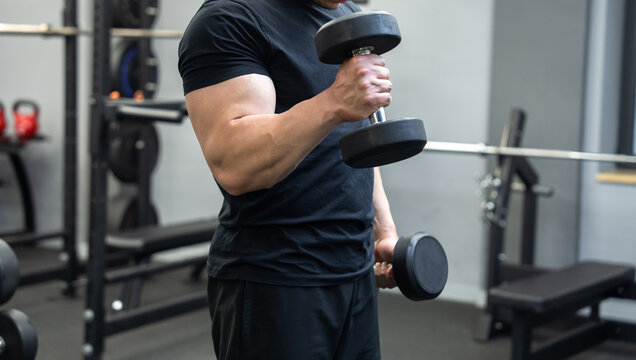 A muscular man is standing in the gym, holding dumbbells, training