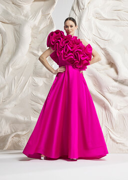 Full length shot of elegant woman wearing luxury magenta pink voluminous ballgown with sculptural 3D floral rosette and train posing gracefully against sculptural white studio wall backdrop