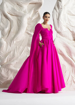 Full length shot of elegant woman wearing luxury fuchsia pink floor-sweeping cape gown with dramatic long train and cowl neck posing gracefully against sculptural white studio wall backdrop