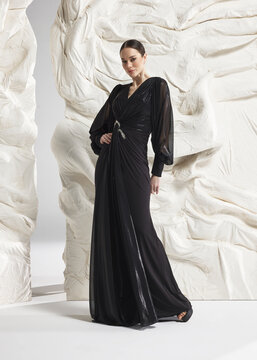 Full length shot of elegant woman wearing luxury black plisse pleated long sleeve column evening gown with cinched waist posing gracefully against sculptural white studio wall backdrop