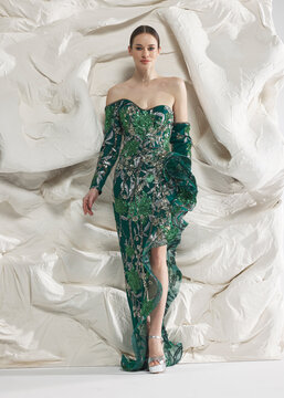 Full length shot of elegant woman wearing luxury emerald green sparkling sequin embroidered evening gown with high leg slit posing gracefully against sculptural white studio wall backdrop