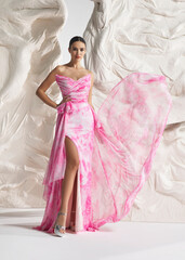 Fototapeta premium Full length shot of elegant woman wearing luxury pink floral watercolor floral organza gown with high-low train and tiered frills posing gracefully against sculptural white studio wall