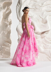 Fototapeta premium Full length shot of elegant woman wearing luxury pink floral flowing watercolor floral gala gown with ruffled train and corset posing gracefully against sculptural white studio wall backdrop