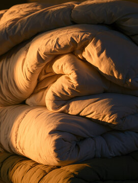 Folding winter blanket stack glowing with golden sunset light, layered quilted comforter fabric creating cozy peaceful home sanctuary with deep shadow and soft texture