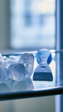 Defrosting scraper beside melting ice chunk inside refrigerator shelf under soft natural window light, featuring cool blue color, reflection, and copy space for commercial stock use