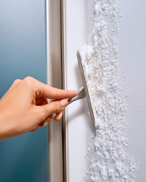 Adult hand scraping freezer frost with metal defrost tool during domestic maintenance in bright light, close view showing realistic skin texture and thick ice buildup