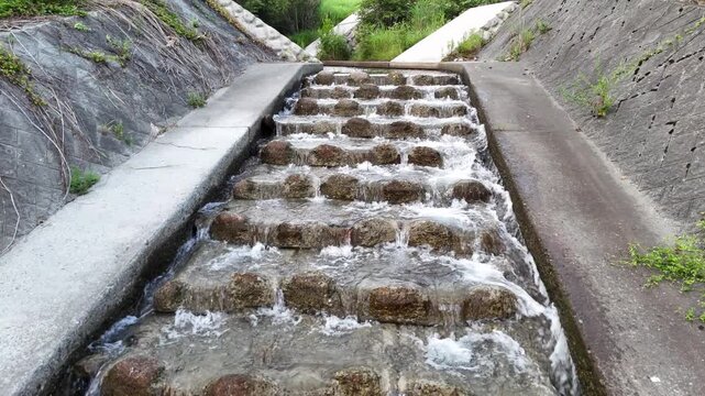 Water flows gently over stepped stone channels. Rustic concrete borders guide the stream's path. Green grass grows between stone and concrete. Natural flow creates soothing, rhythmic motion