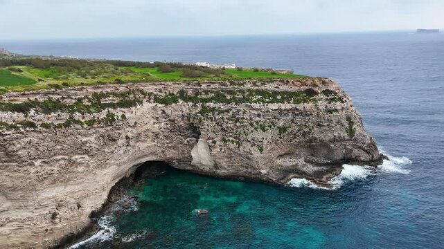 limestone sea cave undercuts cliff face xaqqa cliffs malta aerial drone orbit turquoise cove clifftop farmland rugged headland waves surging distant hazy horizon mediterranean coastline