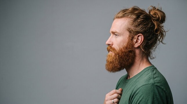 Stylish man with distinctive red beard and hair bun, profile shot on grey background, embodying modern male grooming
