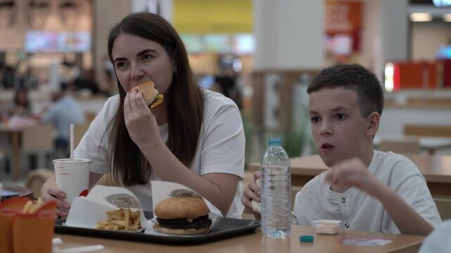 Mother and young son enjoy burgers, fries and wrap at busy food court table, sipping drinks in modern shopping mall setting.