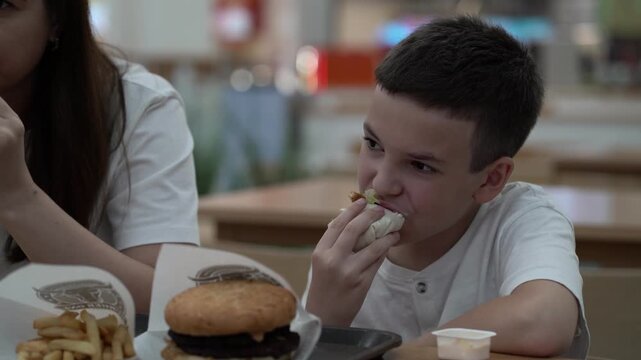 Boy eats Caesar wrap at food court next to mom, with burger and fries on table nearby.
