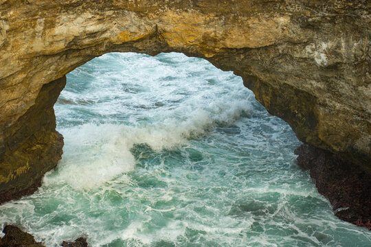 Powerful turquoise ocean waves crashing through the iconic natural stone arch at Karang Bolong Beach in Pacitan. Stunning coastal landscape showcasing sea cave erosion in East Java.