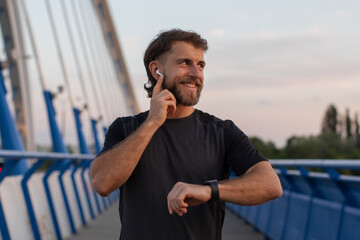 Naklejka premium Smiling sporty man pausing during his run to check time on his smartwatch while standing on modern bridge and touching earphones