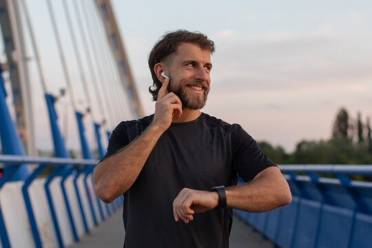 Smiling sporty man pausing during his run to check time on his smartwatch while standing on modern bridge and touching earphones