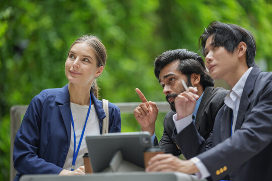 Three colleagues at an outdoor cafe table point at the tablet screen to make a point on a landscaped city plaza surrounded by high rise buildings, takeaway cups crowd the small table top