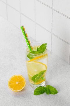 Refreshing homemade lemonade served in clear glass with fresh lemon slices, mint leaves, ice cubes and paper straws on a light kitchen counter against a white tile background with copy space.