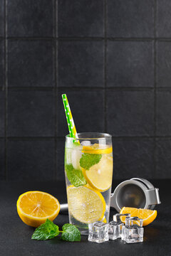 Refreshing homemade lemonade served in clear glass with fresh lemon slices, mint leaves, ice cubes and paper straws on a dark kitchen counter against a black tile background with copy space