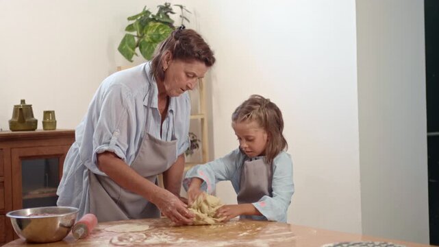 A woman and a girl are making dough together in a kitchen. They mix and knead the dough on a wooden table. Ingredients and tools are present on the table.