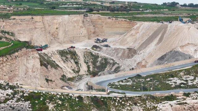 limestone quarry pit cut deep into a malta hillside red dump trucks a tracked excavator work beneath crushing screening machinery a paved road winds down through green farmland framing the rim