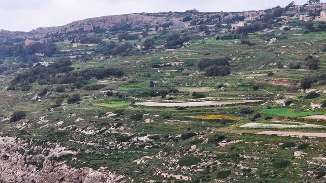 terraced hillside farmland steps down a malta valley in a patchwork of small fields divided by dry rubble walls rugged boulders scatter the foreground a pale stone hilltop village rises on the horizon