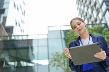 Fototapeta premium A confident businesswoman outdoors reads a document on the tablet in the business district under fresh trees in the public realm of a smart city district, her expression is calm and focused