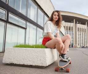 Young woman sitting on a skateboard outside a modern building. Urban youth lifestyle, recreation and street culture concept. © Sergey Chumakov