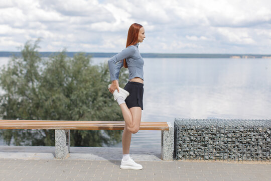 Young woman stretching her quadriceps beside a waterfront before a run. Outdoor warmup, flexibility, running preparation and active lifestyle concept.