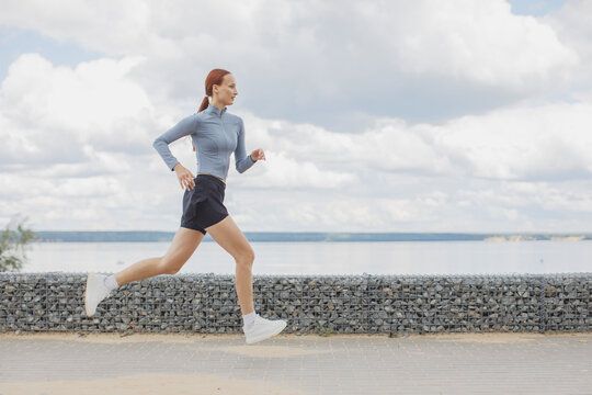 Young woman running along a waterfront promenade under a cloudy sky. Outdoor fitness, jogging and healthy lifestyle concept.