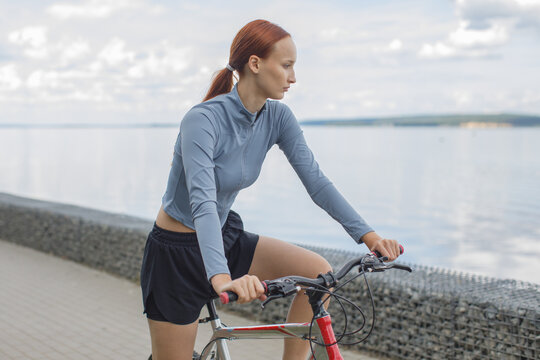 Young woman with a bicycle at the waterfront, preparing for an outdoor workout. Cycling, fitness, recreation and active lifestyle concept.