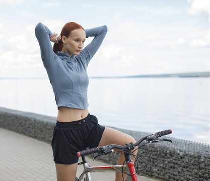 Young woman adjusting her hair beside a bicycle on a waterfront promenade. Cycling preparation, outdoor recreation and active lifestyle concept.
