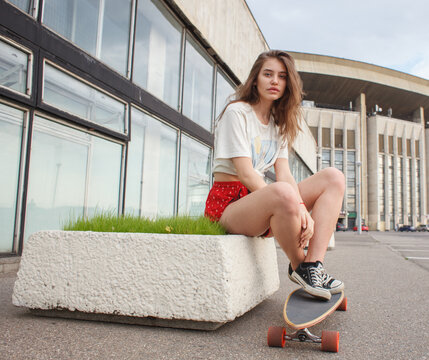 Young woman sitting on a skateboard outside a modern building. Urban youth lifestyle, recreation and street culture concept.