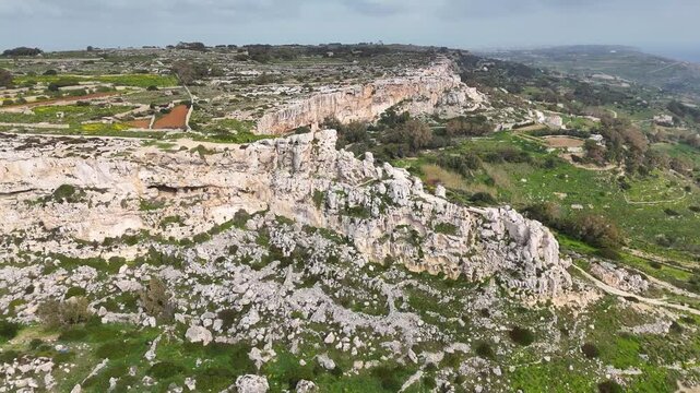 jagged limestone rock field spreads across a karst hillside in malta boulders rubble stacked below a pale cliff band overlooking farmland a distant town on the hazy horizon