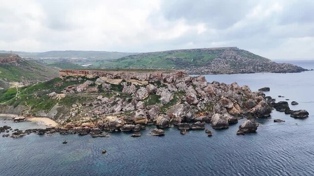 rocky headland lined with sea caves extends into clear blue water off maltas coast reddish clay cliffs pale limestone boulders framing a quiet cove sandy shoreline below green farmland